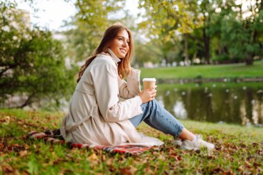 Sonbahar parkında bir bardak sıcak içecekle battaniyede oturan yağmurluk giymiş gülümseyen kadın. Güzel bir kadın göl kenarında eğleniyor. Yürüyüş kavramı, dinlenme.