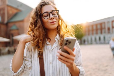 Şehir caddesinde telefonu olan gözlüklü genç bir kadın. Neşeli kadın gün batımında telefonla iletişim kuruyor. Teknoloji kavramı, yürüme. Yaşam biçimi.