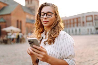 Şehir caddesinde telefonu olan gözlüklü genç bir kadın. Neşeli kadın gün batımında telefonla iletişim kuruyor. Teknoloji kavramı, yürüme. Yaşam biçimi.