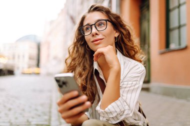 Şehir caddesinde telefonu olan gözlüklü genç bir kadın. Neşeli kadın gün batımında telefonla iletişim kuruyor. Teknoloji kavramı, yürüme. Yaşam biçimi.