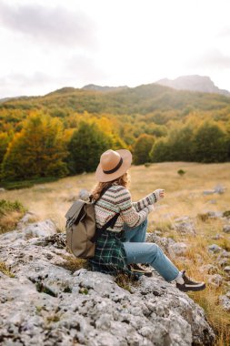 Sırt çantası ve şapkasıyla dağlarda yürüyen genç bir kadının portresi. Gezgin dağ manzarasının tadını çıkarır ve gün batımında özgürlüğü hisseder. Macera, vahşi yaşam konsepti.