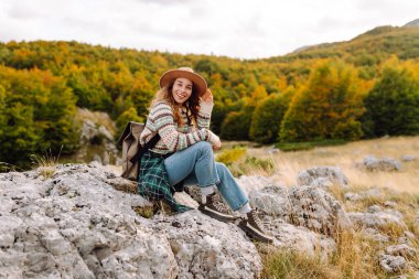Sırt çantası ve şapkasıyla dağlarda yürüyen genç bir kadının portresi. Gezgin dağ manzarasının tadını çıkarır ve gün batımında özgürlüğü hisseder. Macera, vahşi yaşam konsepti.