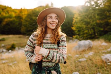 Sırt çantası ve şapkasıyla dağlarda yürüyen genç bir kadının portresi. Gezgin dağ manzarasının tadını çıkarır ve gün batımında özgürlüğü hisseder. Macera, vahşi yaşam konsepti.