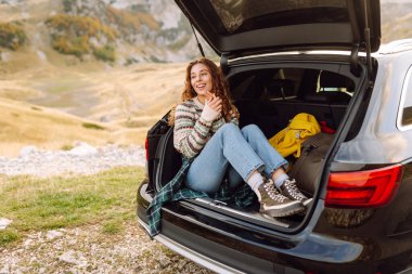 Gülümseyen kadın arabanın bagajında oturuyor ve güneşli bir günde dağ manzarasının tadını çıkarıyor. Yolculuktaki güzel bayan yürüyüşçü özgürlüğü hissediyor. Doğa ve özgürlük kavramı.
