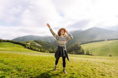 Rahat süveter ve şapkalı neşeli kadın çimenli dağlık arazide oturuyor. Yeşil tepelerdeki gezgin özgürlüğü hissediyor ve dağ manzarasının tadını çıkarıyor. Etkin yaşam biçimi.
