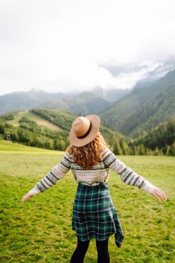 Rahat süveter ve şapkalı neşeli kadın çimenli dağlık arazide oturuyor. Yeşil tepelerdeki gezgin özgürlüğü hissediyor ve dağ manzarasının tadını çıkarıyor. Etkin yaşam biçimi.