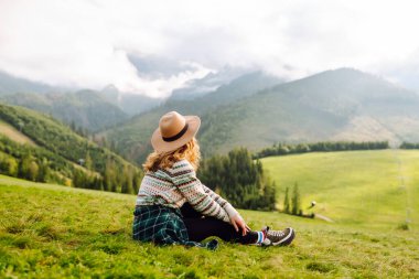 Rahat süveter ve şapkalı neşeli kadın çimenli dağlık arazide oturuyor. Yeşil tepelerdeki gezgin özgürlüğü hissediyor ve dağ manzarasının tadını çıkarıyor. Etkin yaşam biçimi.