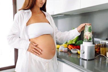 Güzel hamile kadın güneşli bir günde sıcak mutfakta yeşil smoothie hazırlıyor. Bebek bekleyen tatlı bir kadın diyetini izliyor ve smoothie içiyor. Yemek pişirme, sağlık, kolay hamilelik kavramı. Yaşam biçimi.