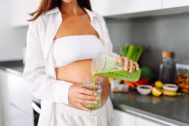 Güzel hamile kadın güneşli bir günde sıcak mutfakta yeşil smoothie hazırlıyor. Bebek bekleyen tatlı bir kadın diyetini izliyor ve smoothie içiyor. Yemek pişirme, sağlık, kolay hamilelik kavramı. Yaşam biçimi.