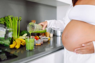 Güzel hamile kadın güneşli bir günde sıcak mutfakta yeşil smoothie hazırlıyor. Bebek bekleyen tatlı bir kadın diyetini izliyor ve smoothie içiyor. Yemek pişirme, sağlık, kolay hamilelik kavramı. Yaşam biçimi.
