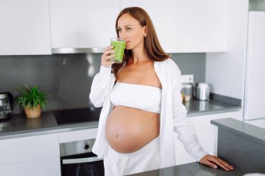 Güzel hamile kadın güneşli bir günde sıcak mutfakta yeşil smoothie hazırlıyor. Bebek bekleyen tatlı bir kadın diyetini izliyor ve smoothie içiyor. Yemek pişirme, sağlık, kolay hamilelik kavramı. Yaşam biçimi.