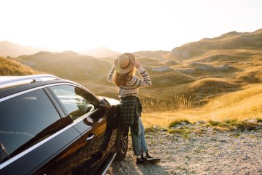 Şapka ve kazak giyen genç bayan gezgin gün batımında araba yakınında dağ manzarasının tadını çıkarıyor. Güzel bir kadın araba yolculuğunda özgürlüğünü hisseder. Macera, ulaşım kavramı.