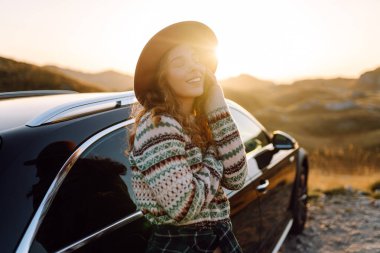 Şapka ve kazak giyen genç bayan gezgin gün batımında araba yakınında dağ manzarasının tadını çıkarıyor. Güzel bir kadın araba yolculuğunda özgürlüğünü hisseder. Macera, ulaşım kavramı.