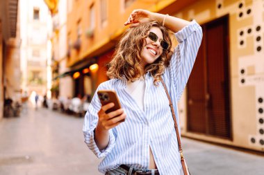 Güneş gözlüklü, elinde telefonla Avrupa sokaklarında yürüyen neşeli bir kadın. Bir turist telefonla konuşuyor ve güneşli bir günün tadını çıkarıyor. Turizm ve macera konsepti. Yaşam biçimi.