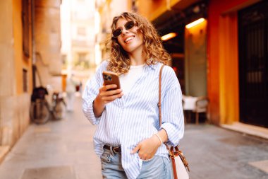 Güneş gözlüklü, elinde telefonla Avrupa sokaklarında yürüyen neşeli bir kadın. Bir turist telefonla konuşuyor ve güneşli bir günün tadını çıkarıyor. Turizm ve macera konsepti. Yaşam biçimi.