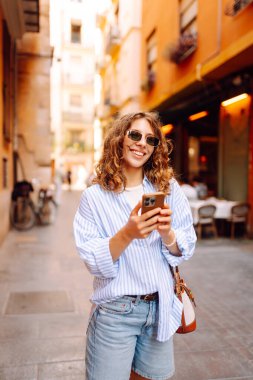 Güneş gözlüklü, elinde telefonla Avrupa sokaklarında yürüyen neşeli bir kadın. Bir turist telefonla konuşuyor ve güneşli bir günün tadını çıkarıyor. Turizm ve macera konsepti. Yaşam biçimi.