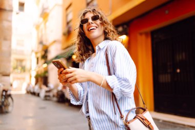 Güneş gözlüklü, elinde telefonla Avrupa sokaklarında yürüyen neşeli bir kadın. Bir turist telefonla konuşuyor ve güneşli bir günün tadını çıkarıyor. Turizm ve macera konsepti. Yaşam biçimi.