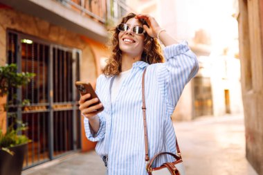 Güneş gözlüklü, elinde telefonla Avrupa sokaklarında yürüyen neşeli bir kadın. Bir turist telefonla konuşuyor ve güneşli bir günün tadını çıkarıyor. Turizm ve macera konsepti. Yaşam biçimi.