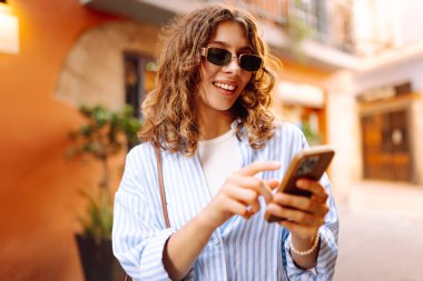 Güneş gözlüklü, elinde telefonla Avrupa sokaklarında yürüyen neşeli bir kadın. Bir turist telefonla konuşuyor ve güneşli bir günün tadını çıkarıyor. Turizm ve macera konsepti. Yaşam biçimi.