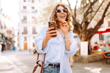 Güneş gözlüklü, elinde telefonla Avrupa sokaklarında yürüyen neşeli bir kadın. Bir turist telefonla konuşuyor ve güneşli bir günün tadını çıkarıyor. Turizm ve macera konsepti. Yaşam biçimi.