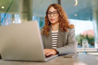 Gözlüklü güzel bir kadın güneşli bir günde bir terasta otururken dizüstü bilgisayarda çalışıyor. Diz üstü bilgisayarı olan serbest çalışan bir kadın açık havada rahat bir atmosferin tadını çıkarıyor. İş ve teknoloji kavramı.