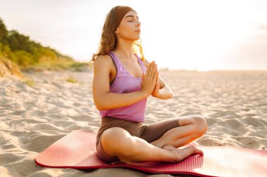 Genç bir kadın, plajda güneşin altında yoga pozu veriyor. Egzersiz yapan atletik bir kadın ve deniz manzarasının tadını çıkarıyor. Yoga, rahatlama, meditasyon kavramı.