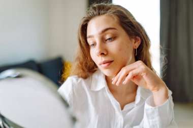 Genç bir kadının portresi küçük bir aynaya bakıyor ve rahat bir odada cilt kusurlarını inceliyor. Şirin bir kadın elleriyle içeride kızarıklıklara dokunuyor. Cilt bakımı, cilt bakımı kavramı.