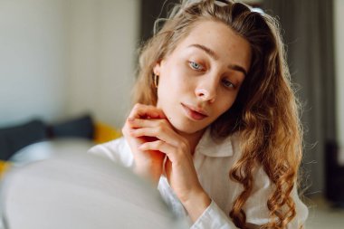 Genç bir kadının portresi küçük bir aynaya bakıyor ve rahat bir odada cilt kusurlarını inceliyor. Şirin bir kadın elleriyle içeride kızarıklıklara dokunuyor. Cilt bakımı, cilt bakımı kavramı.