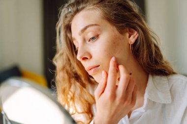 Genç bir kadının portresi küçük bir aynaya bakıyor ve rahat bir odada cilt kusurlarını inceliyor. Şirin bir kadın elleriyle içeride kızarıklıklara dokunuyor. Cilt bakımı, cilt bakımı kavramı.