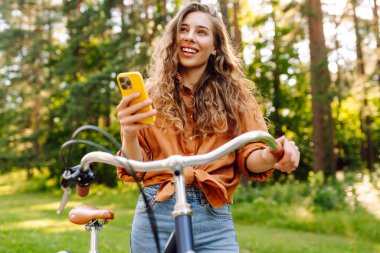 Kıvırcık saçlı ve telefonlu neşeli kadın portresi güneşli parkta bisiklete biniyor. Neşeli kadın gün batımında bisiklet sürmekten hoşlanır. Yürüme, teknoloji konsepti.