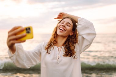 Telefonlu mutlu kadın gün batımında kumlu sahilde selfie çekiyor. Deniz manzarası seyreden ve dışarıda blog yazan genç bir kadın. Teknoloji, blog yazma ve doğa kavramı.