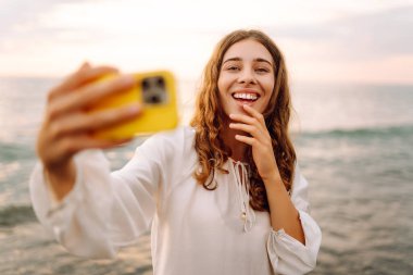 Telefonlu mutlu kadın gün batımında kumlu sahilde selfie çekiyor. Deniz manzarası seyreden ve dışarıda blog yazan genç bir kadın. Teknoloji, blog yazma ve doğa kavramı.