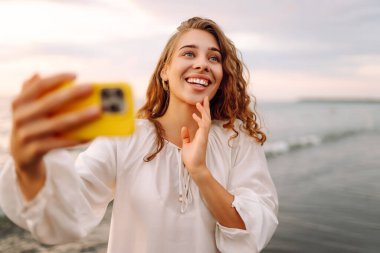 Telefonlu mutlu kadın gün batımında kumlu sahilde selfie çekiyor. Deniz manzarası seyreden ve dışarıda blog yazan genç bir kadın. Teknoloji, blog yazma ve doğa kavramı.