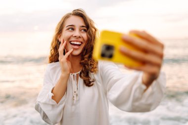 Telefonlu mutlu kadın gün batımında kumlu sahilde selfie çekiyor. Deniz manzarası seyreden ve dışarıda blog yazan genç bir kadın. Teknoloji, blog yazma ve doğa kavramı.