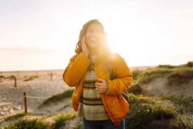 Sırt çantası ve parlak ceketi olan bir bayan gezgin gün batımında kumlu bir sahilde yürümekten zevk alır. Mutlu kadın açık havada özgürlüğü hissediyor. Doğa, macera konsepti.