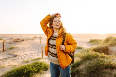 Sırt çantası ve parlak ceketi olan bir bayan gezgin gün batımında kumlu bir sahilde yürümekten zevk alır. Mutlu kadın açık havada özgürlüğü hissediyor. Doğa, macera konsepti.