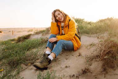 Sırt çantası ve parlak ceketi olan bir bayan gezgin gün batımında kumlu bir sahilde yürümekten zevk alır. Mutlu kadın açık havada özgürlüğü hissediyor. Doğa, macera konsepti.