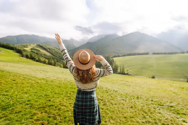 Rahat süveter ve şapkalı neşeli kadın çimenli dağlık arazide oturuyor. Yeşil tepelerdeki gezgin özgürlüğü hissediyor ve dağ manzarasının tadını çıkarıyor. Etkin yaşam biçimi.