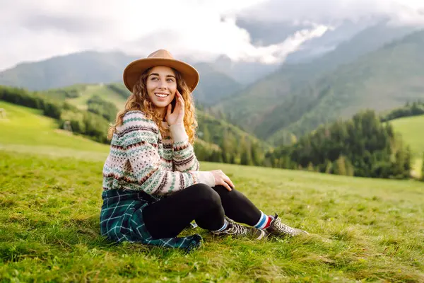 Rahat süveter ve şapkalı neşeli kadın çimenli dağlık arazide oturuyor. Yeşil tepelerdeki gezgin özgürlüğü hissediyor ve dağ manzarasının tadını çıkarıyor. Etkin yaşam biçimi.