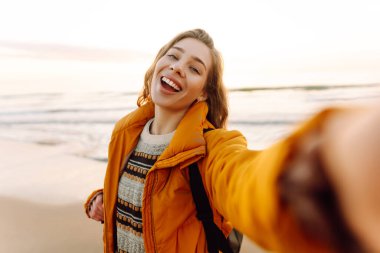 Gün batımında sahilde yürürken selfie çeken parlak ceketli kadın portresi. Telefon blogları olan güzel bir gezgin, doğanın tadını çıkarıyor. Bloglama, eğlenceli konsept.