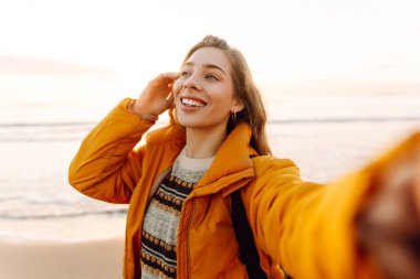 Gün batımında sahilde yürürken selfie çeken parlak ceketli kadın portresi. Telefon blogları olan güzel bir gezgin, doğanın tadını çıkarıyor. Bloglama, eğlenceli konsept.