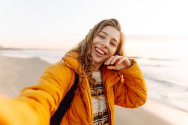 Gün batımında sahilde yürürken selfie çeken parlak ceketli kadın portresi. Telefon blogları olan güzel bir gezgin, doğanın tadını çıkarıyor. Bloglama, eğlenceli konsept.