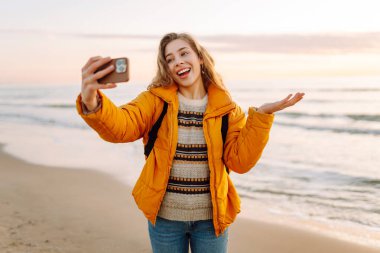 Gün batımında sahilde yürürken selfie çeken parlak ceketli kadın portresi. Telefon blogları olan güzel bir gezgin, doğanın tadını çıkarıyor. Bloglama, eğlenceli konsept.