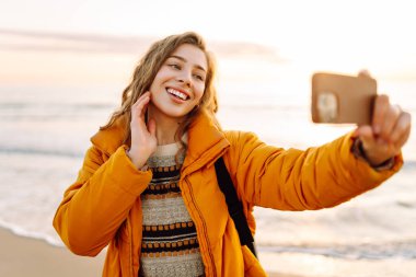 Gün batımında sahilde yürürken selfie çeken parlak ceketli kadın portresi. Telefon blogları olan güzel bir gezgin, doğanın tadını çıkarıyor. Bloglama, eğlenceli konsept.
