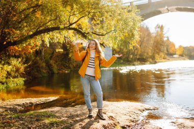 Mutlu kadın, sonbaharda elinde bir telefonla nehir kıyısında duruyor. Bir turist günbatımı ormanında su kenarında yürürken selfie çekiyor. Hafta sonu konsepti, blog yazarı.