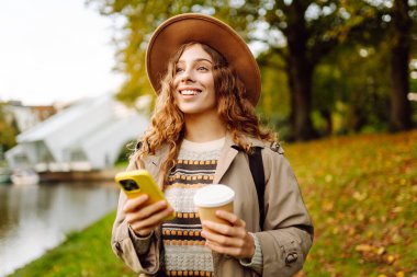 Bir sonbahar parkında yürürken elinde bir fincan sıcak içecek ve telefon tutan şapkalı genç bir kadının portresi. Neşeli turist blogları, kahve içmek ve güneşli bir günün tadını çıkarmak. Yaşam biçimi.