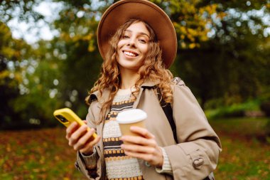 Bir sonbahar parkında yürürken elinde bir fincan sıcak içecek ve telefon tutan şapkalı genç bir kadının portresi. Neşeli turist blogları, kahve içmek ve güneşli bir günün tadını çıkarmak. Yaşam biçimi.