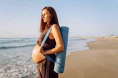 Güneşte sahilde elinde yoga minderiyle gülümseyen hamile kadın. Doğanın tadını çıkaran bir çocuk ve deniz kenarında yürüyüş bekleyen genç bir kadın. Rahatlama, yürüyüş konsepti.