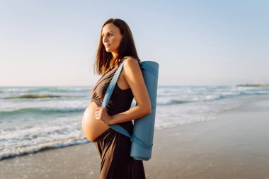 Güneşte sahilde elinde yoga minderiyle gülümseyen hamile kadın. Doğanın tadını çıkaran bir çocuk ve deniz kenarında yürüyüş bekleyen genç bir kadın. Rahatlama, yürüyüş konsepti.