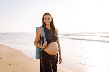Güneşte sahilde elinde yoga minderiyle gülümseyen hamile kadın. Doğanın tadını çıkaran bir çocuk ve deniz kenarında yürüyüş bekleyen genç bir kadın. Rahatlama, yürüyüş konsepti.